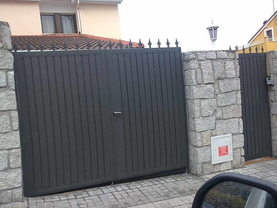 Puerta gris y pequeña puerta a juego en muro de piedra con una casa al fondo.