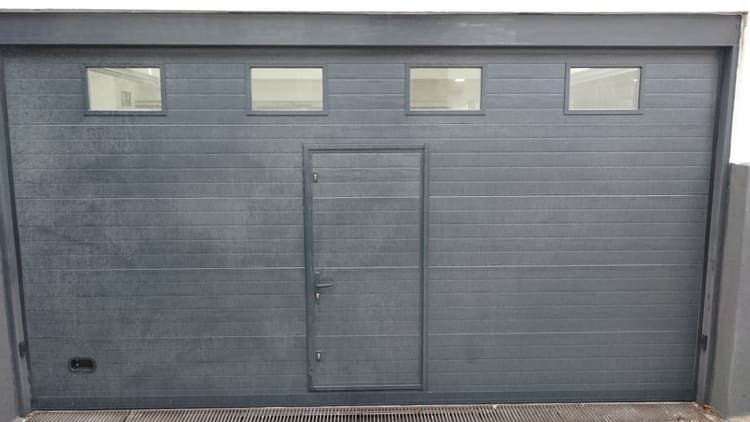 Puerta de garaje gris con cuatro pequeñas ventanas y puerta peatonal.