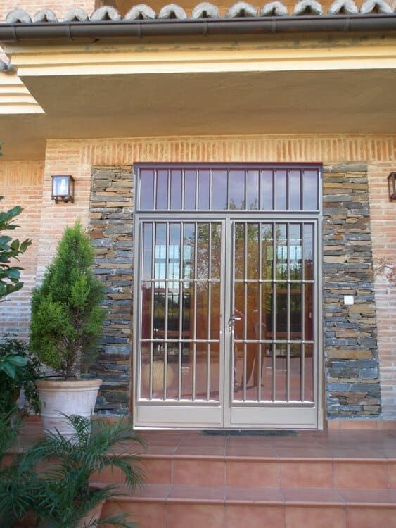 Puertas dobles de vidrio con rejas de seguridad, enmarcadas por piedra y ladrillo exterior, debajo de una entrada cubierta.