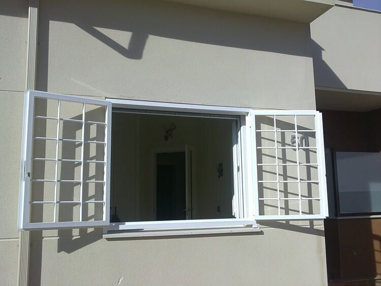 Ventana con marco blanco y rejas abiertas en un edificio beige, que revela una puerta interior.