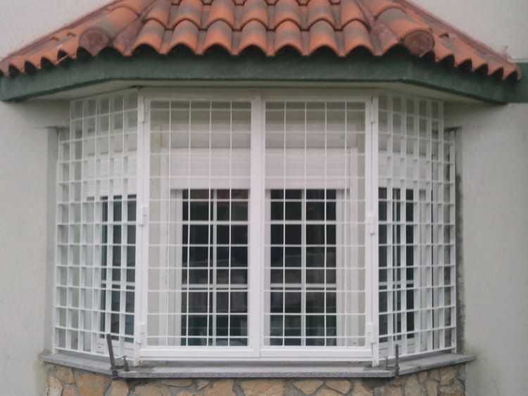 Rejas de seguridad con rejilla blanca en un ventanal con techo de tejas de terracota arriba y base de piedra abajo.
