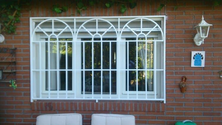 Ventana con barrotes blancos sobre pared de ladrillo, con tres elementos decorativos arqueados.