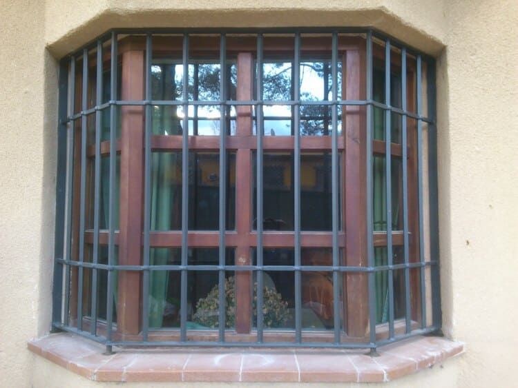 Ventana con rejas de metal oscuro y marco de madera en un edificio de estuco.