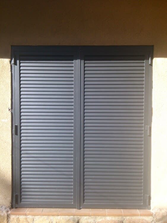 Persianas grises cerradas que cubren una ventana. Instaladas en un edificio marrón claro.