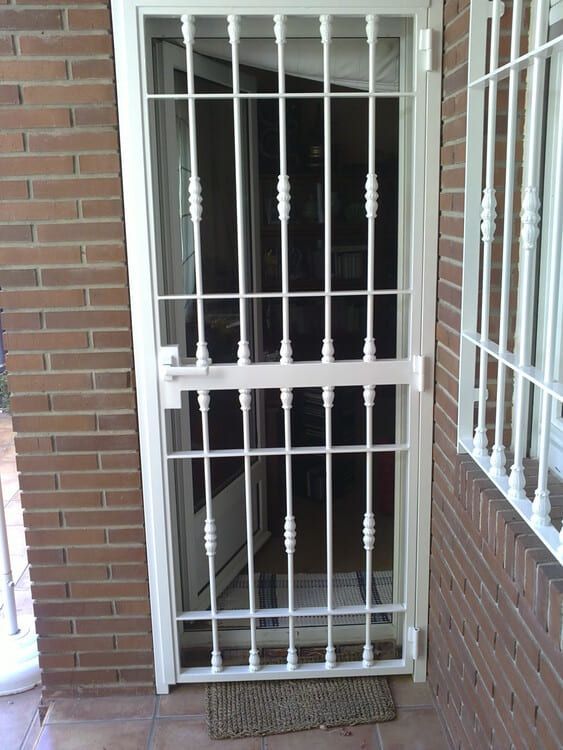 Puerta de seguridad blanca con rejas sobre puerta de cristal, situada en un edificio de ladrillo.