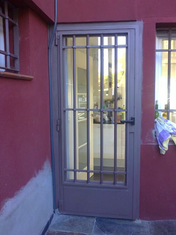 Puerta de seguridad marrón con barras verticales y panel de vidrio, ubicada en un exterior de edificio rojo.