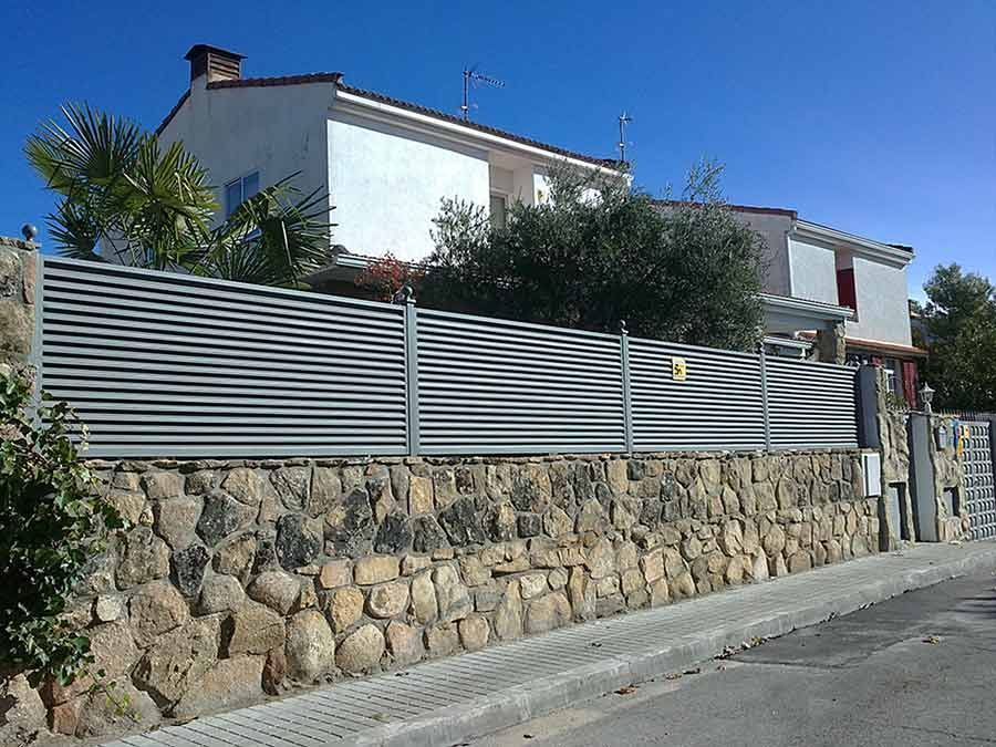 Muro de piedra rematado con una valla metálica horizontal frente a una casa blanca en un día soleado.