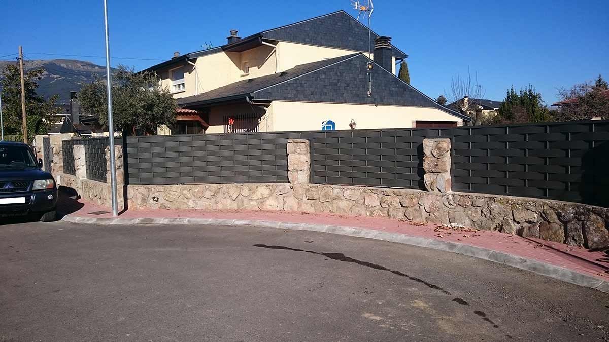 Muro de piedra y valla frente a una casa en la calle. Día soleado, cielo azul.