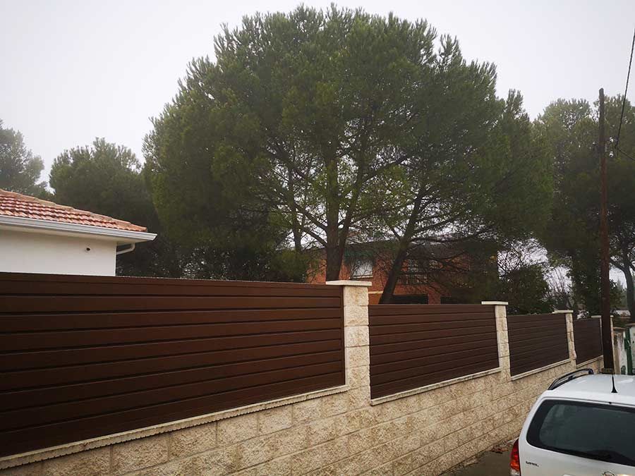 Valla horizontal de listones marrones con base de piedra beige, pinos altos y una casa. Cielo nublado.