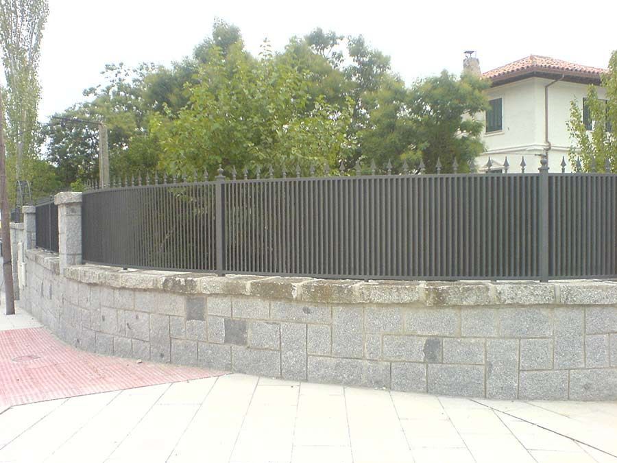 Valla de metal gris sobre un muro de piedra, árboles al fondo y acera al frente.