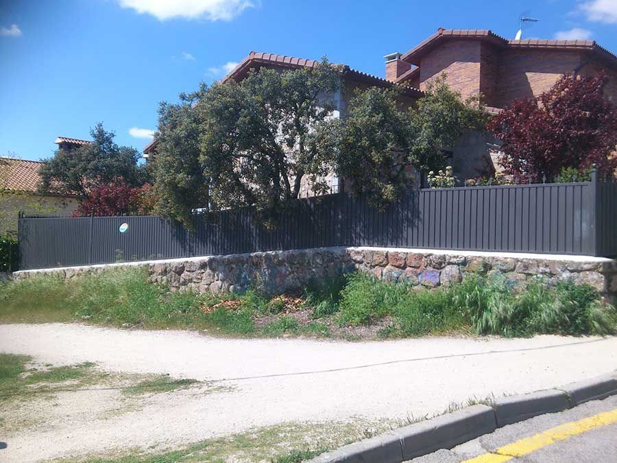 Una casa con un muro de piedra y una valla gris, árboles y arbustos frente a un cielo azul.