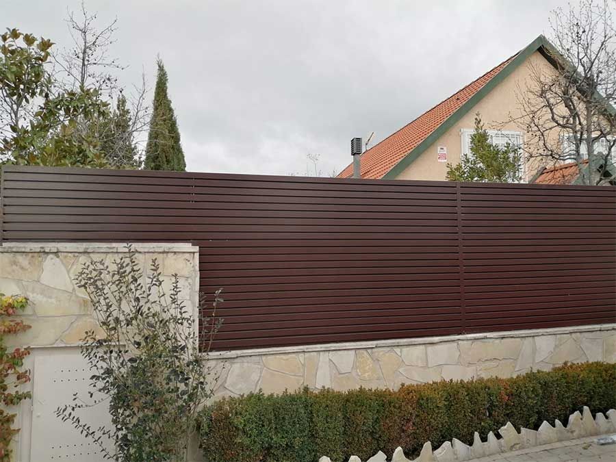 Valla de listones horizontales de color marrón sobre un muro de piedra, frente a una casa con techo rojo.