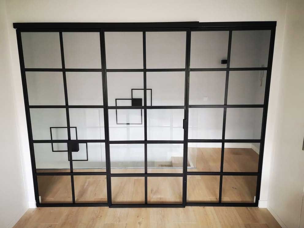 Puertas corredizas de vidrio con marco negro y diseño de cuadrícula geométrica; instaladas en una habitación con piso de madera clara.