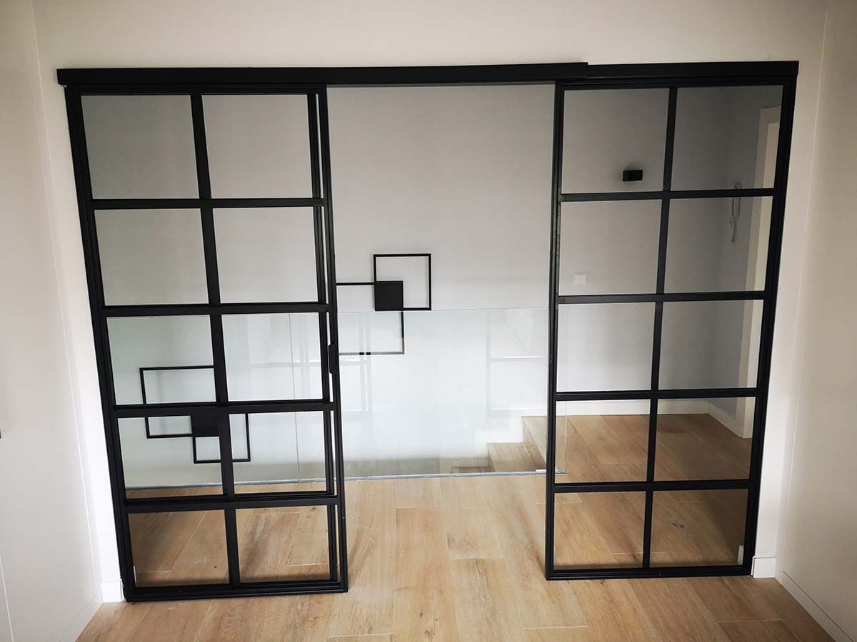Puertas corredizas de vidrio con marco de metal negro, abiertas, en un interior moderno con escaleras y piso de madera.