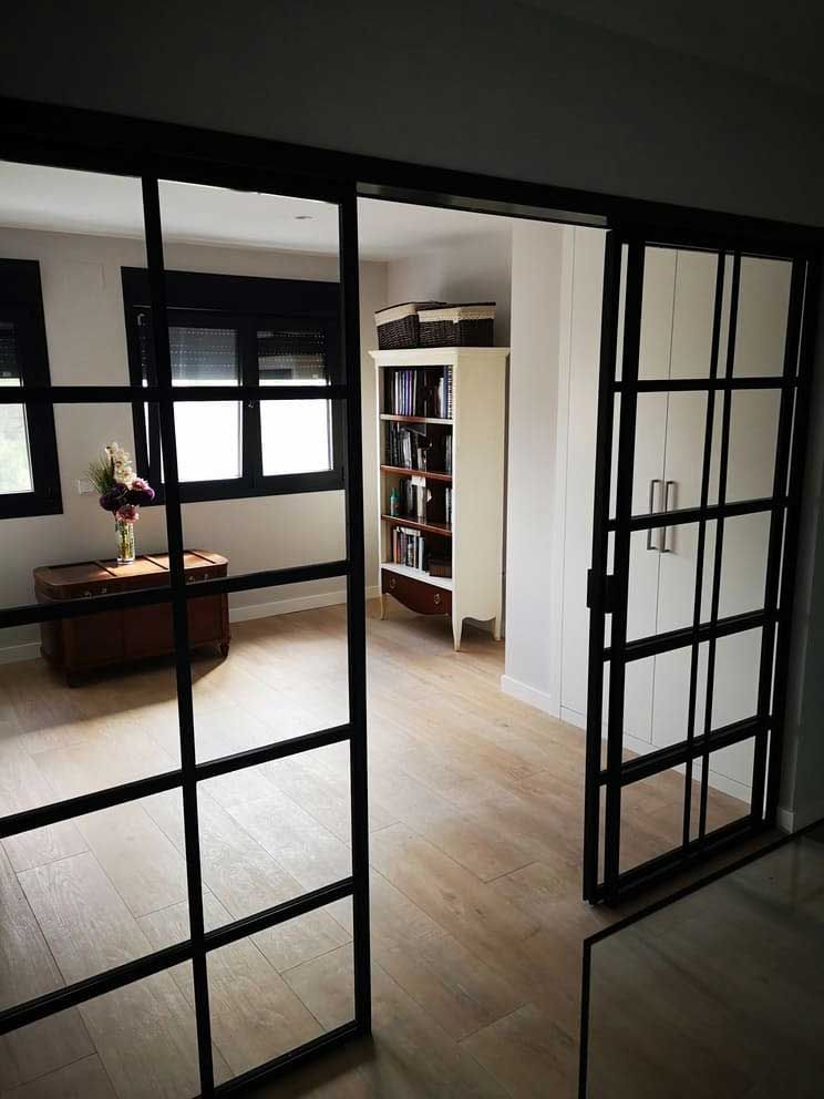 Las puertas corredizas de vidrio con marcos de metal negro se abren a una habitación con pisos de madera clara, una estantería y una ventana.