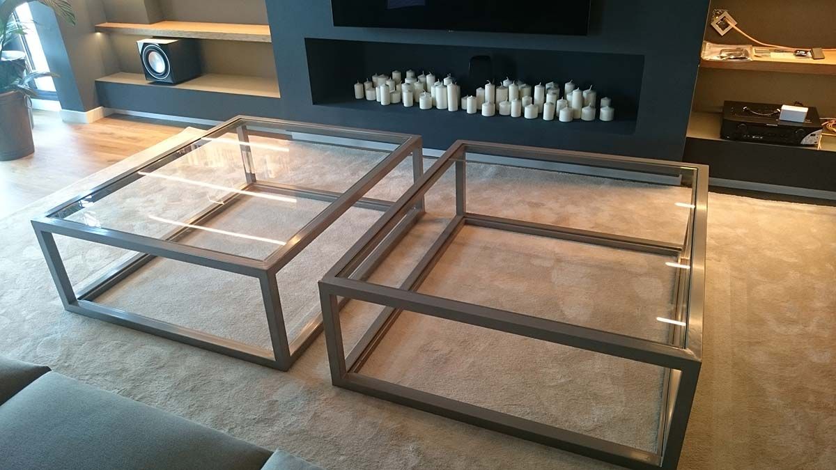 Dos mesas de centro con tapa de cristal y marcos de metal oscuro sobre una alfombra clara, cerca de una chimenea y estantes.