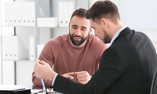 Zwei Männer in einem Büro, einer im Anzug, der andere im Pullover, untersuchen etwas aufmerksam.