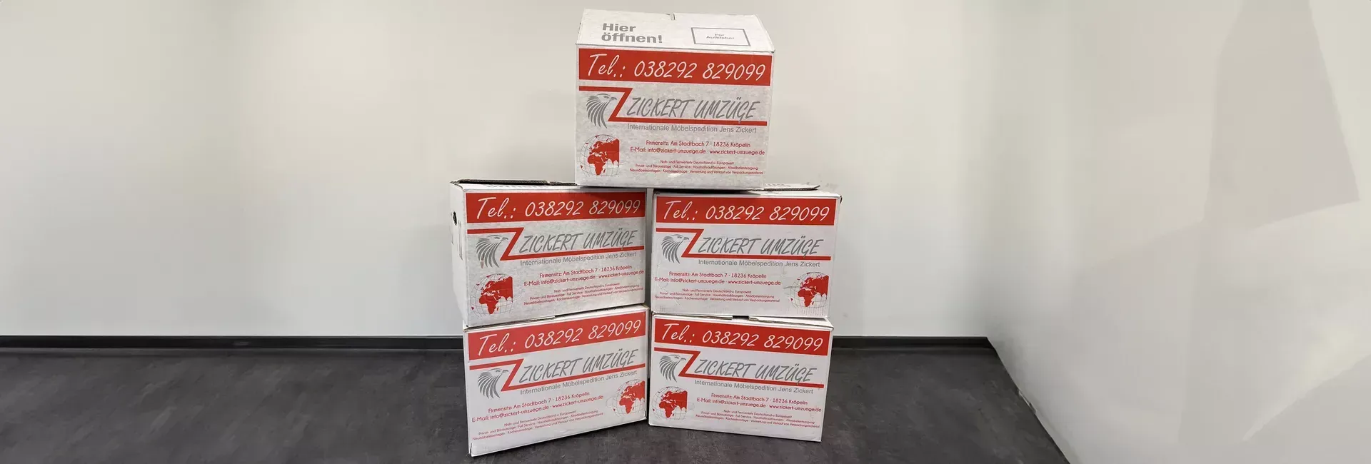 Fünf rot-weiße Kisten sind pyramidenförmig auf einer dunklen Fläche vor einer weißen Wand gestapelt.