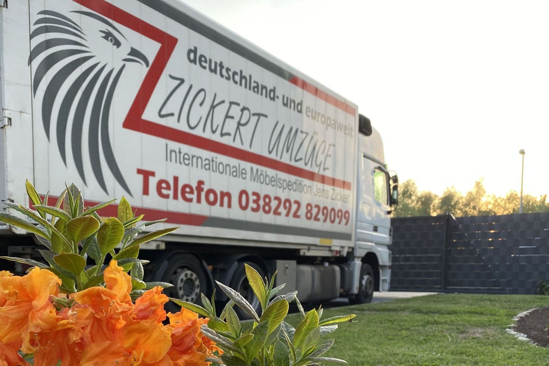 Umzugswagen mit Firmenlogo und Kontaktinformationen, geparkt neben einem grünen Rasen mit orangefarbenen Blumen.
