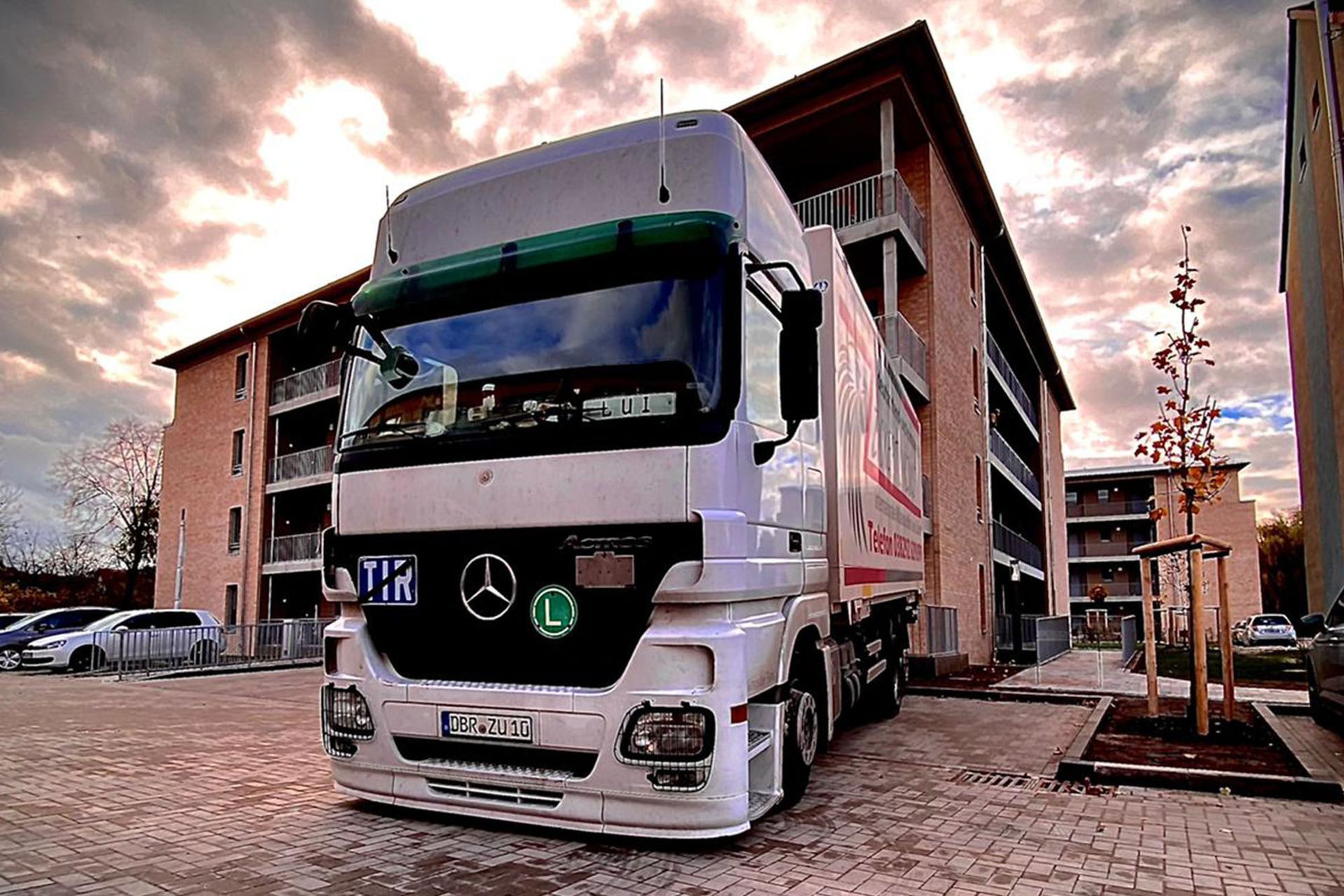 Weißer Sattelschlepper parkt vor einem hellen Gebäude. Über ihm ist ein bewölkter Himmel.