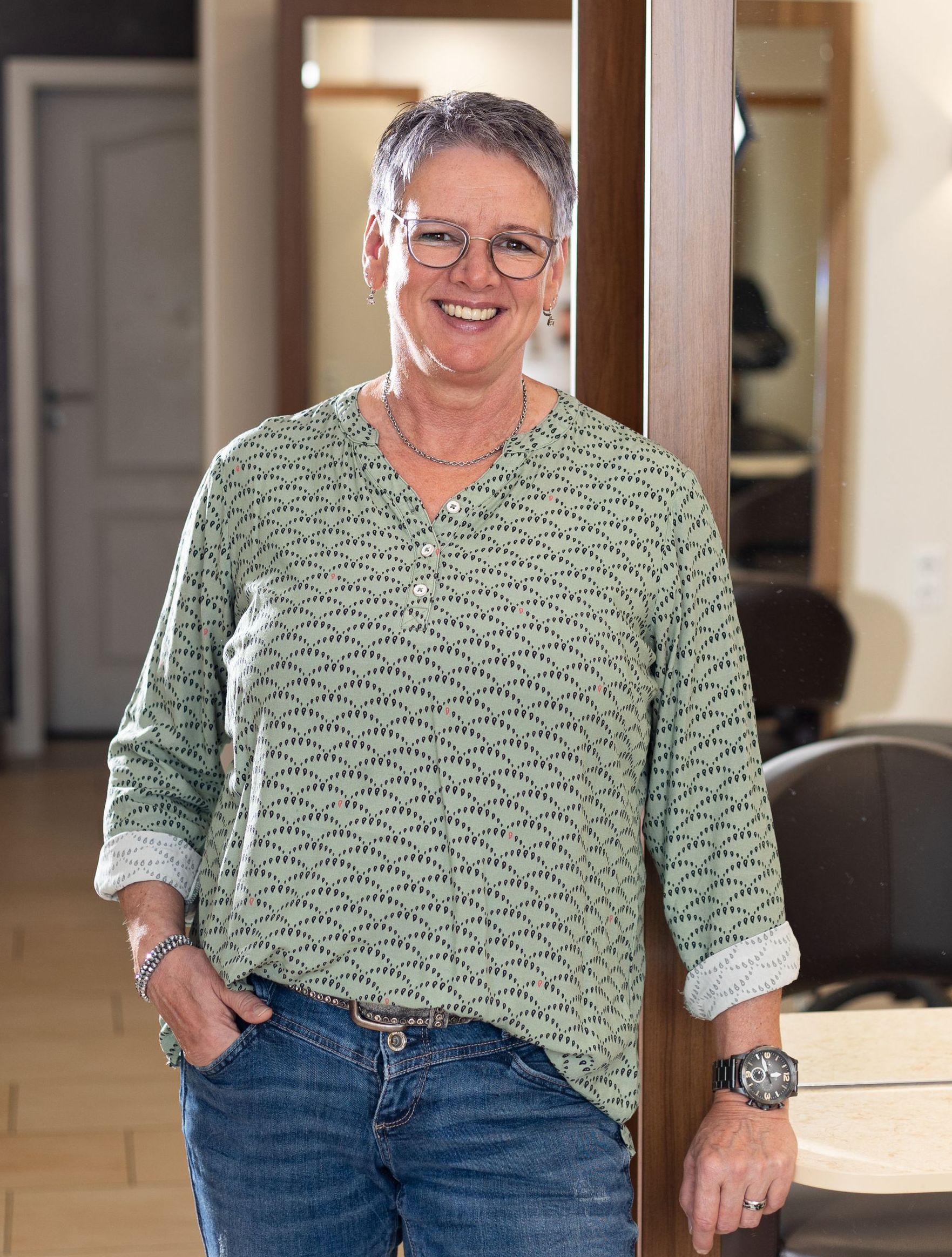 Eine Frau in einem grünen Hemd und Jeans steht mit den Händen in den Taschen in einem Raum.