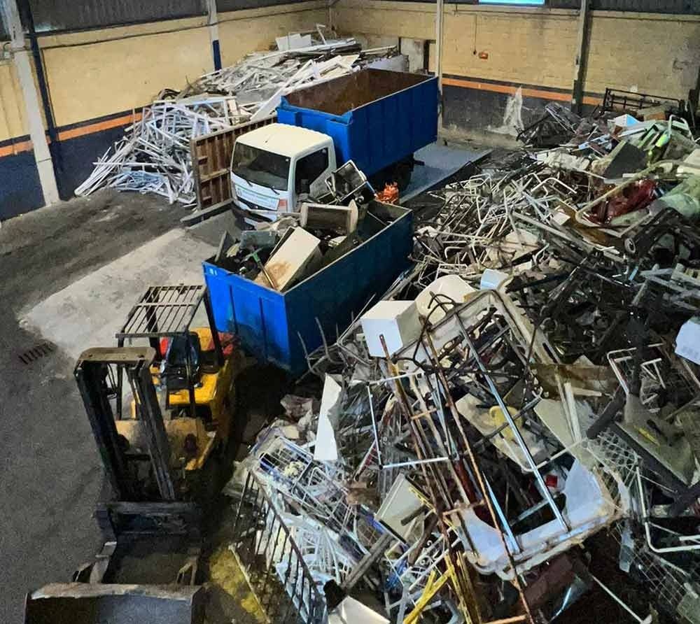 Una carretilla elevadora carga chatarra en contenedores azules en un patio industrial desordenado.