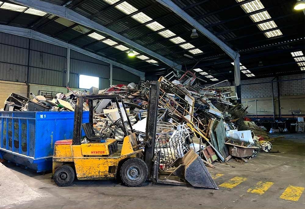 Una carretilla elevadora amarilla carga chatarra en un contenedor azul dentro de un almacén industrial.