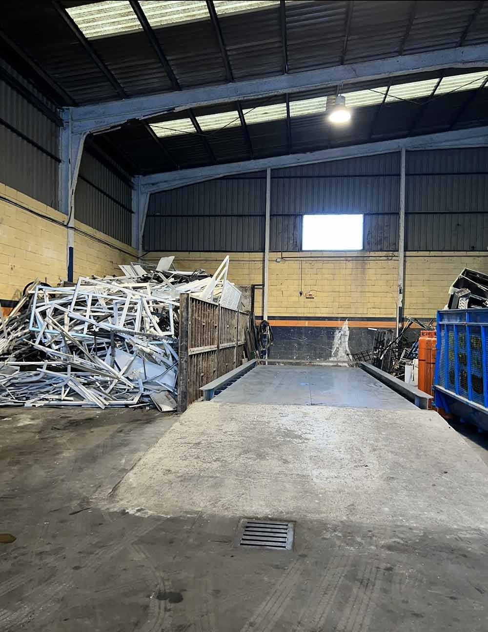 Interior de una nave industrial con contenedores para chatarra y suelo de hormigón bajo un alto techo metálico.