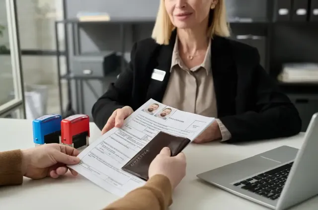 Persona entregando pasaporte y documentos a una mujer en un escritorio con una computadora portátil y sellos.