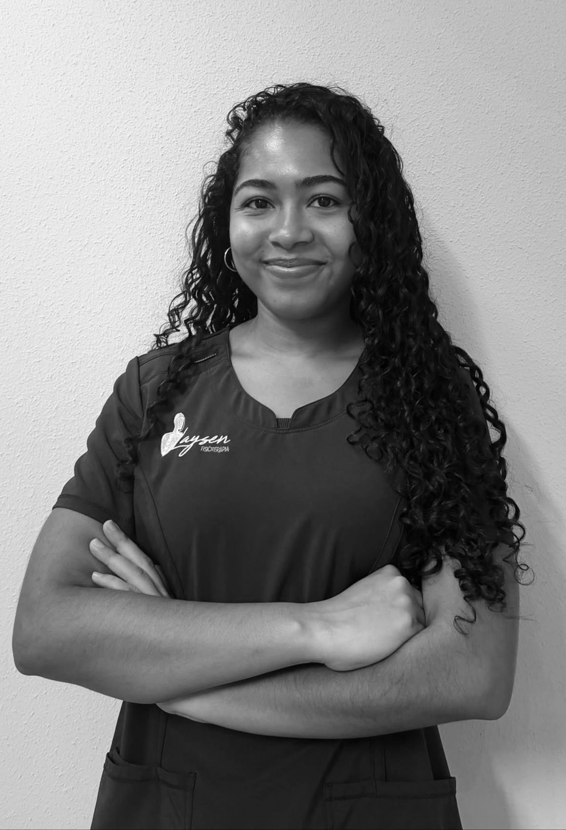 Persona sonriente con gafas y blusa oscura, de pie con los brazos cruzados sobre un fondo liso.