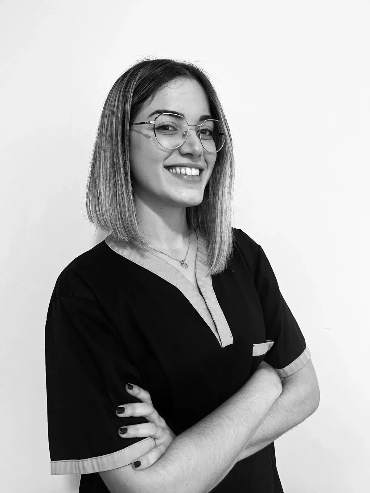 Persona sonriente con gafas y blusa oscura, de pie con los brazos cruzados sobre un fondo liso.