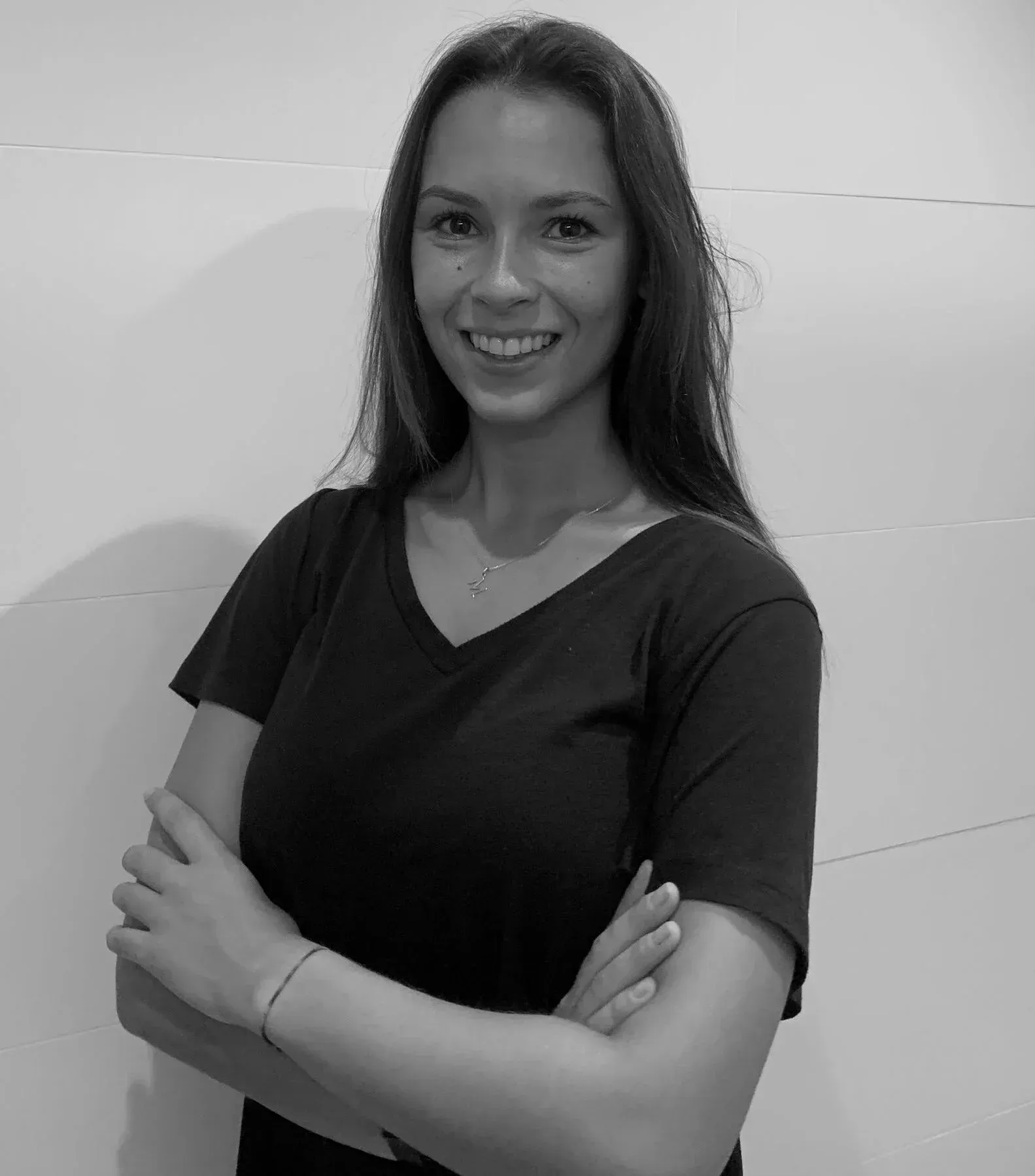 Mujer sonriente con una camiseta oscura de pie con los brazos cruzados contra una pared lisa.