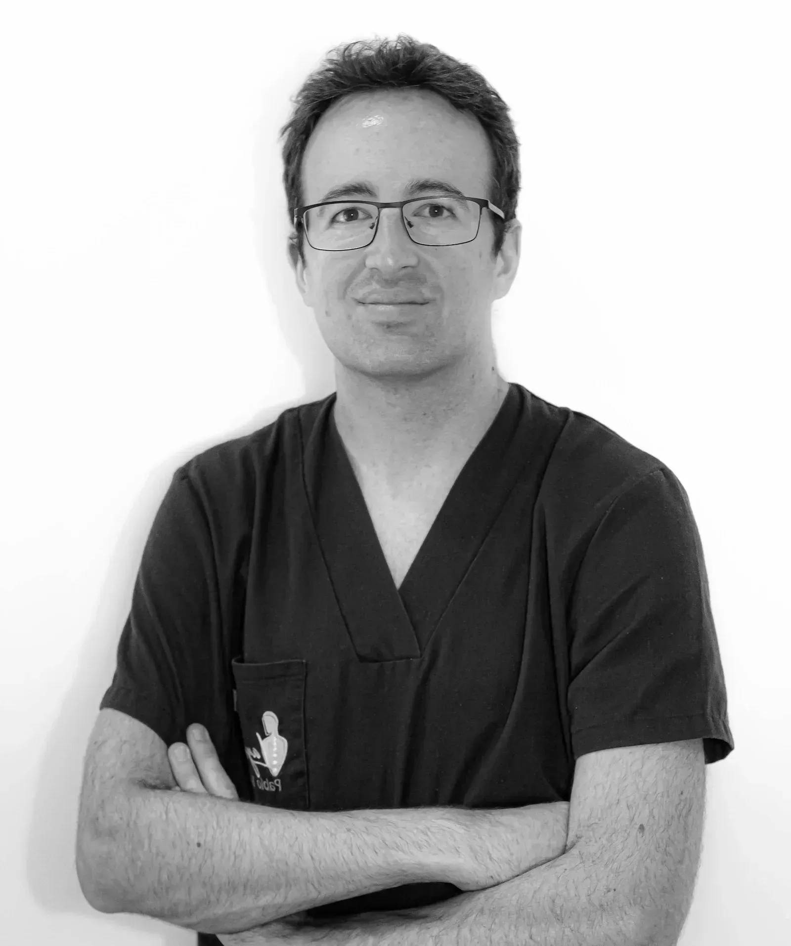 Persona con uniforme médico oscuro y gafas, con los brazos cruzados, de pie sobre un fondo claro liso.
