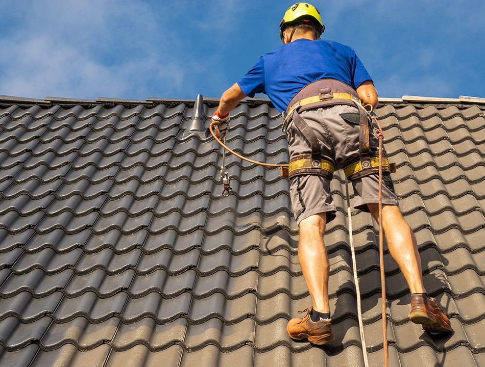 Un ouvrier raccordé à une ligne de vie sur une toiture projette de la peinture sur les tuiles à l'aide d'un pulvérisateur