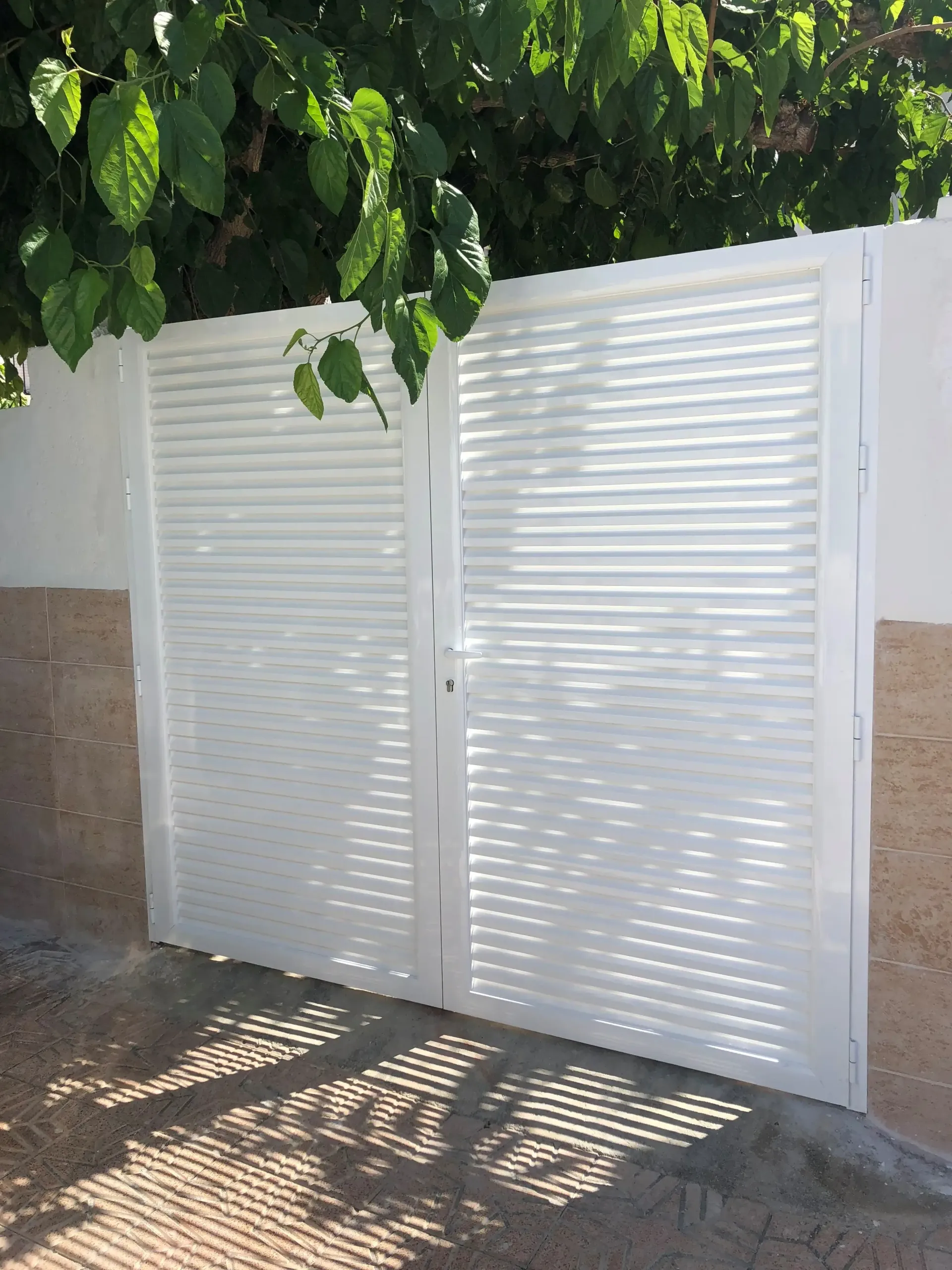 Una verja de metal blanco con paneles de celosía se encuentra junto a una pared de color beige, a la sombra de las hojas de los árboles.