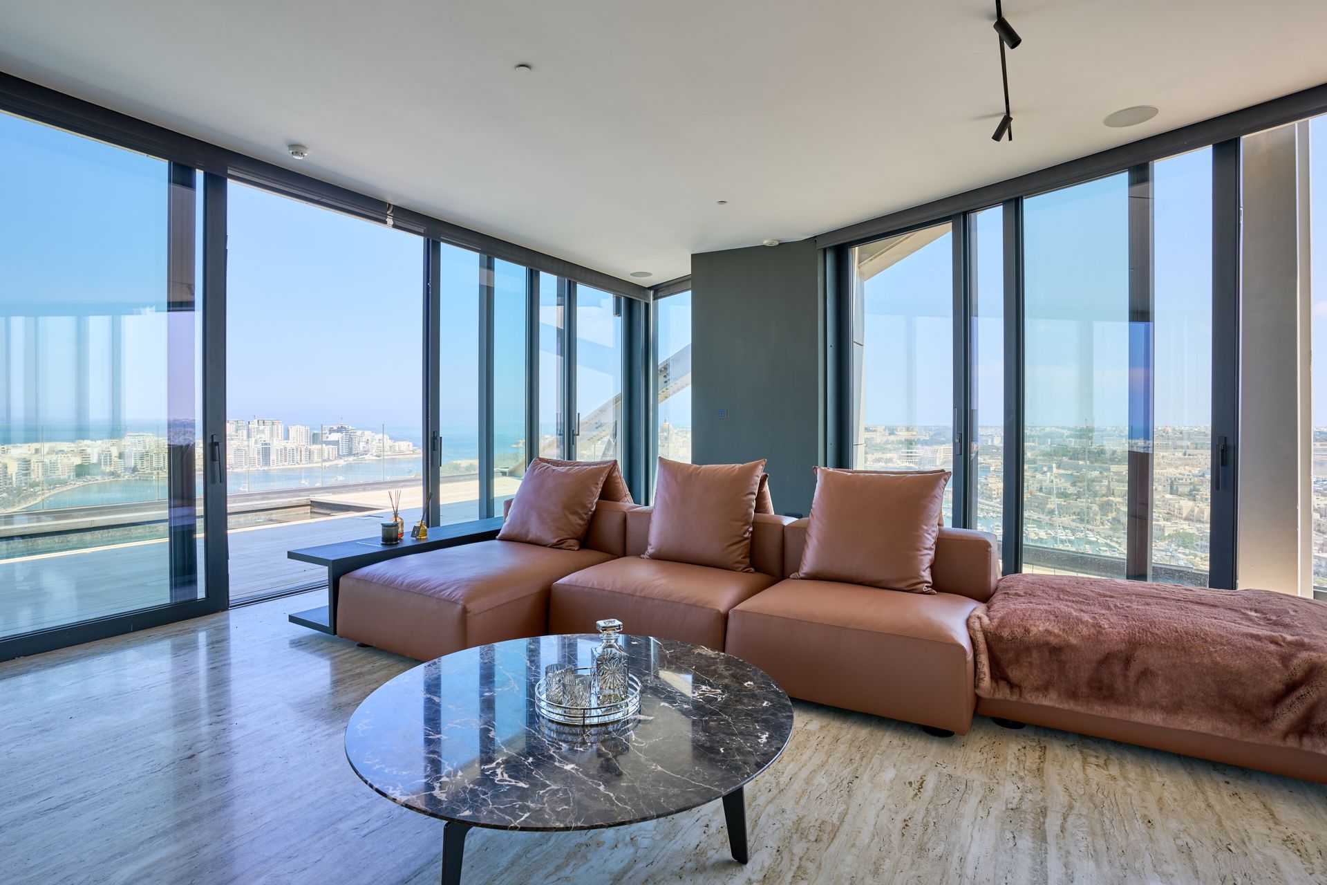 Modern living room with tan sectional sofa, glass coffee table, and floor-to-ceiling windows overlooking the city.