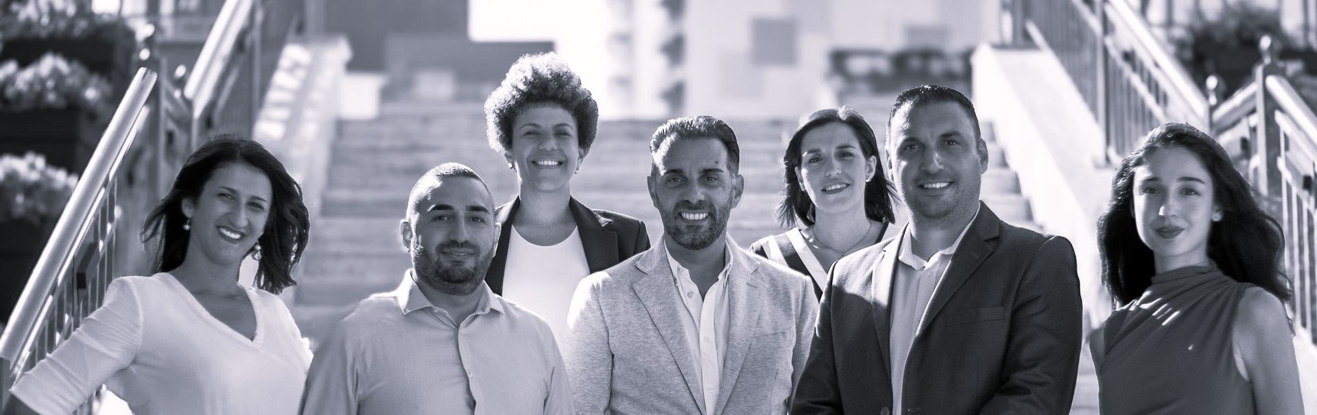 Six smiling coworkers standing on outdoor stairs, posing together in business attire