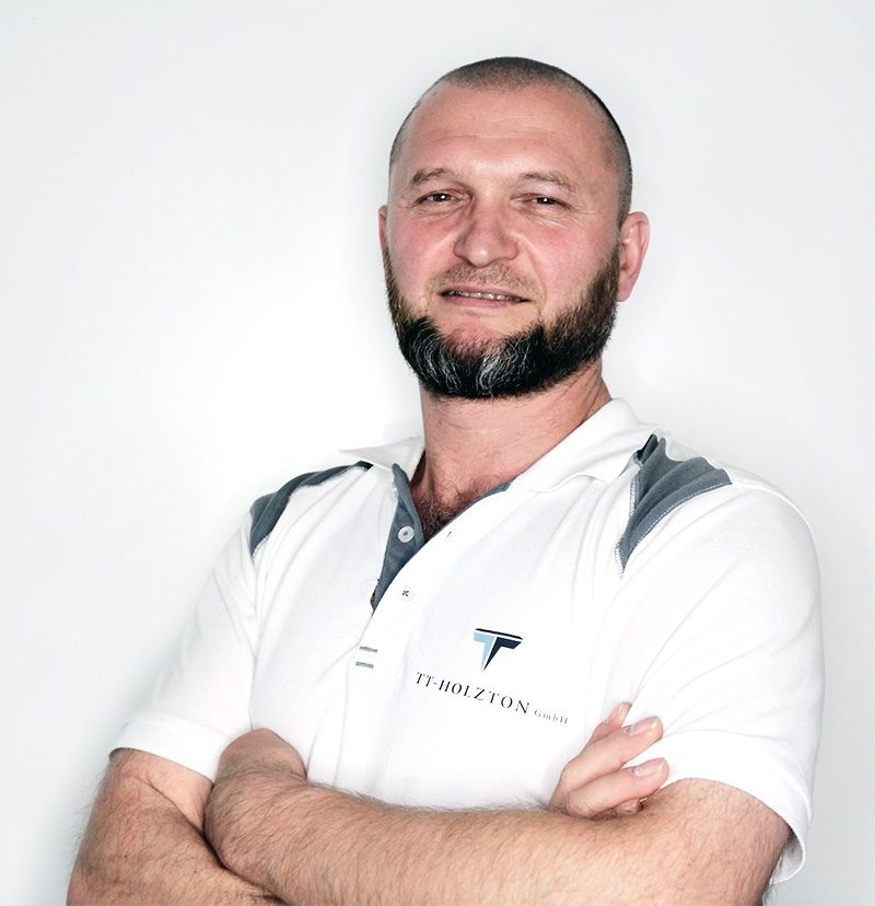 Mann mit Bart und verschränkten Armen, der ein weisses Poloshirt trägt, lächelt in die Kamera.