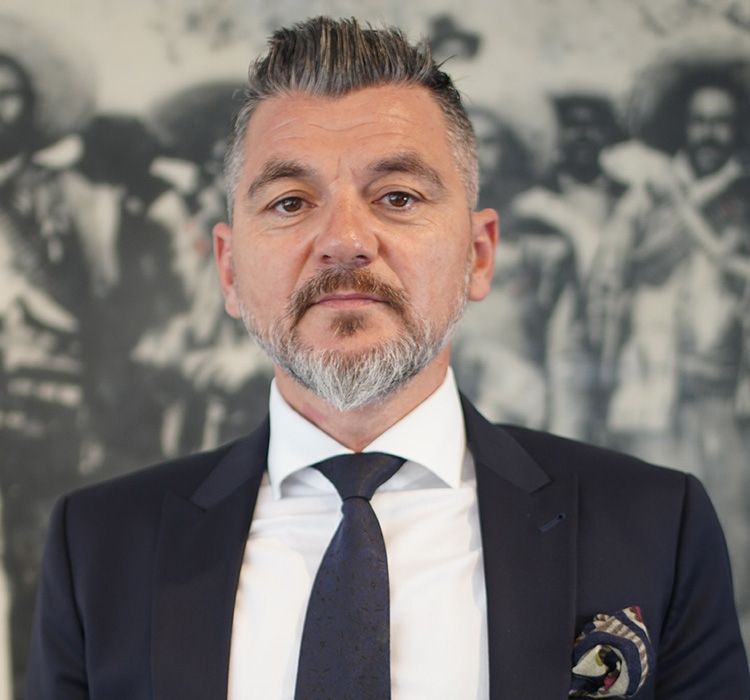 Ein Mann im Anzug blickt mit neutralem Gesichtsausdruck in die Kamera. Im Hintergrund ist ein historisches Schwarz-Weiss-Foto zu sehen.