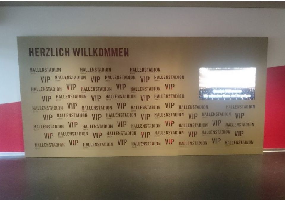 Goldfarbenes Willkommensschild mit dem Text „HERZLICH WILLKOMMEN“, wiederholtem „VIP“, „WALLSTADION“ und einem beleuchteten Bildschirm.