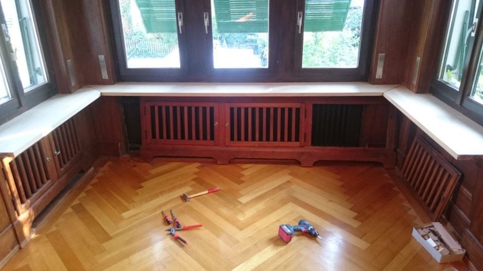 Holzgetäfelter Raum mit Heizkörperverkleidung unterhalb der Fenster, Fischgrätparkett, verstreutes Werkzeug, Tageslicht fällt hindurch.
