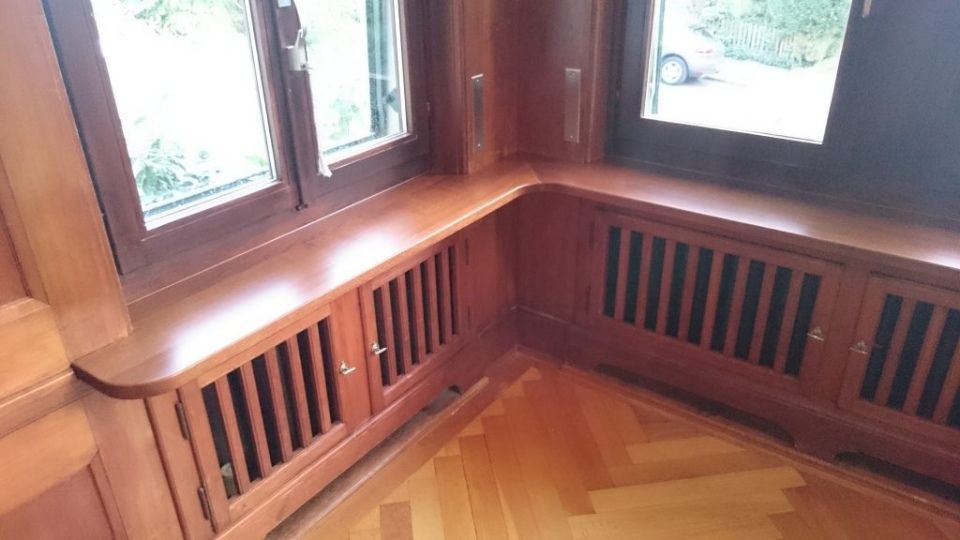Holzfensterbank mit eingebauten Schränken und Lamellenlüftung, in einer Ecke neben Fenstern und Parkettboden.