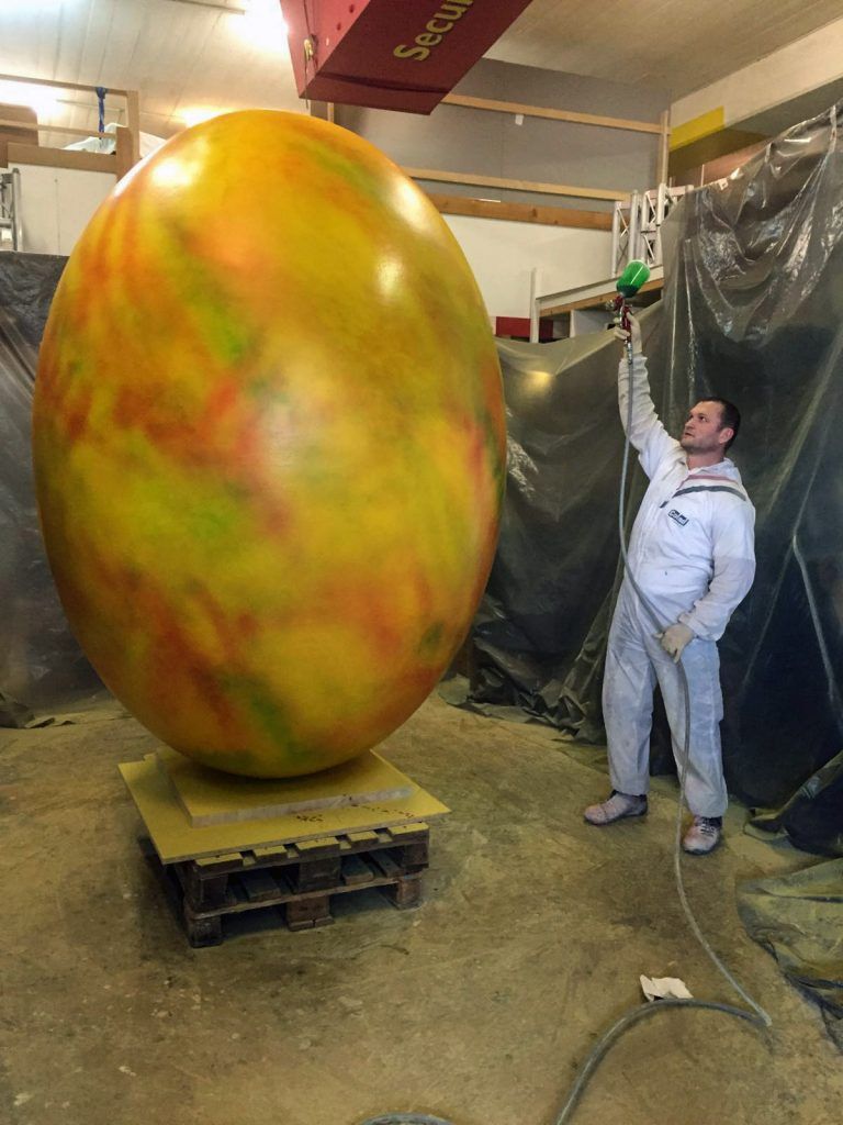 Ein Mann besprüht ein riesiges, buntes Ei mit Farbe. Die Szene spielt in einer Werkstatt, in der gelbe Farbspritzer zu sehen sind.