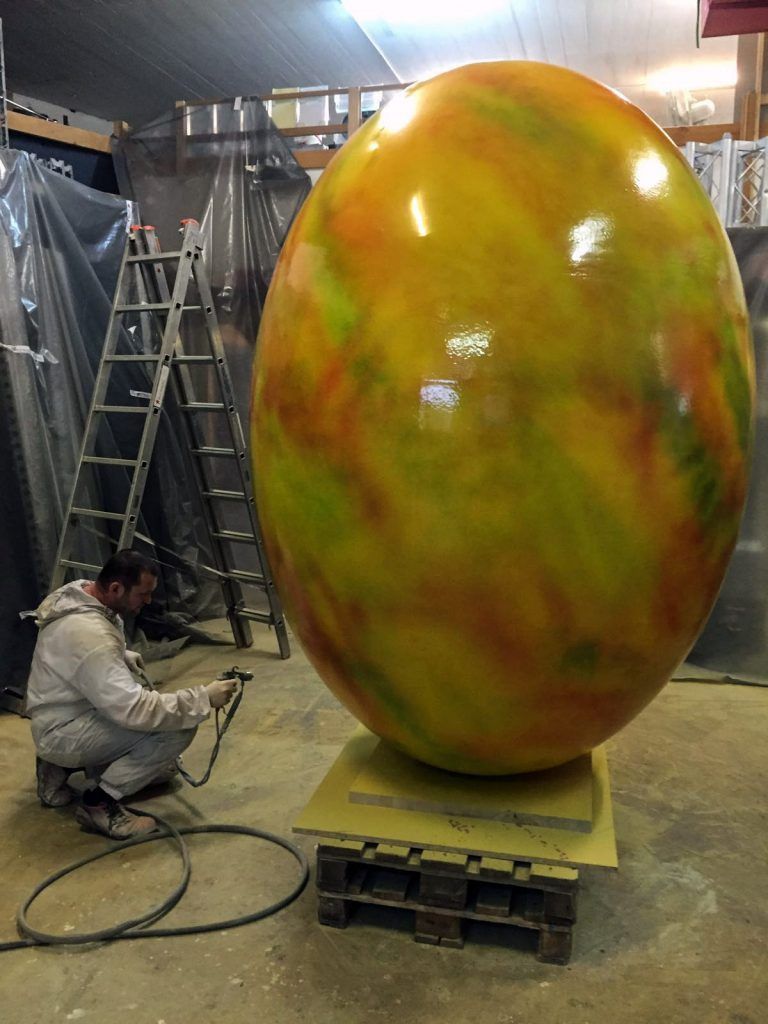 Eine Person besprüht in einer Werkstatt eine grosse, eiförmige Skulptur mit gelben, grünen und orangen Wirbeln.