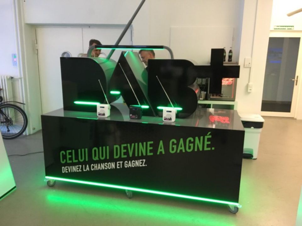 Schwarzer Tresen mit beleuchteten Buchstaben „NAB+“, grünen Lichtern, französischem Text und zwei sichtbaren Personen.