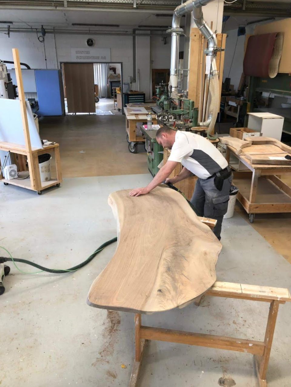 Ein Mann schleift in einer Werkstatt eine grosse, unregelmässige Holzplatte. Ein Werkstattsauger ist angeschlossen.
