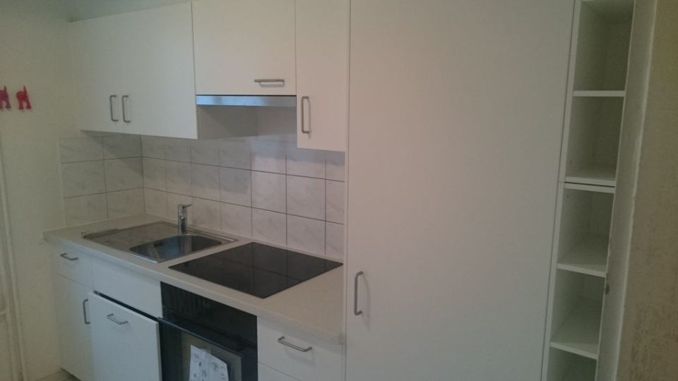 Kleine weisse Küche mit Herd, Spüle, Schränken und hohem Stauraum.