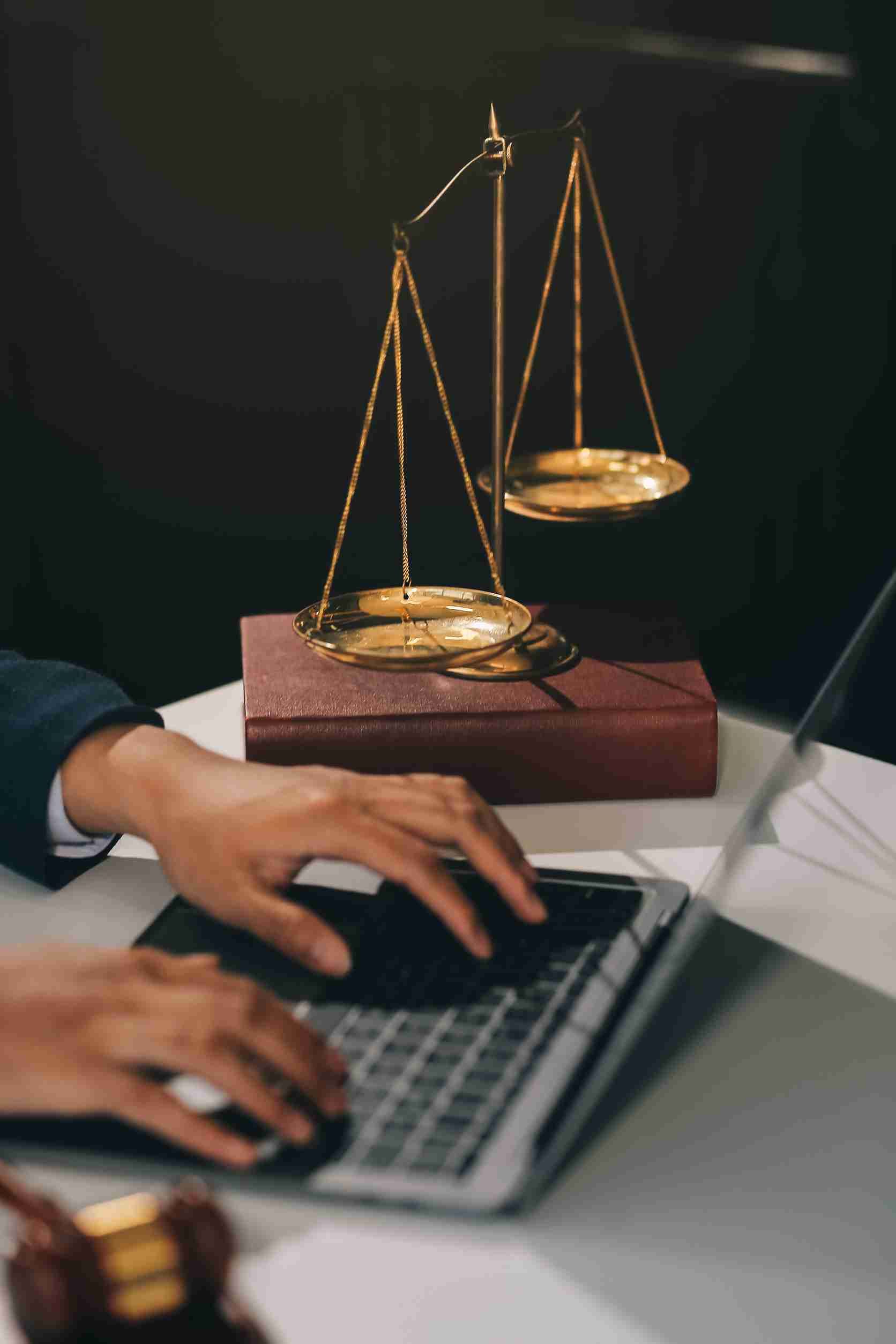Person typing on a laptop with golden scales of justice and a gavel in the foreground on a white table.