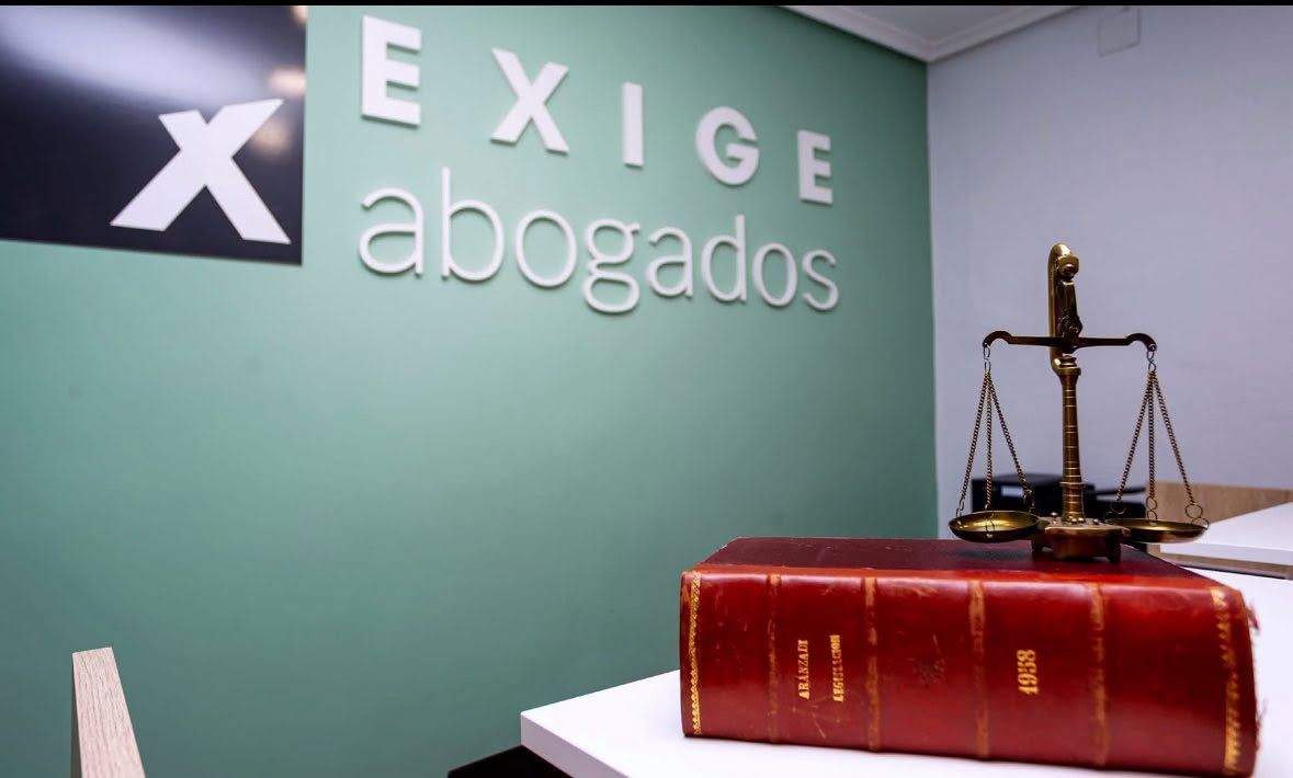 Letrero de la oficina de “EXIGE abogados” con una balanza legal y un libro de leyes rojo sobre un escritorio.