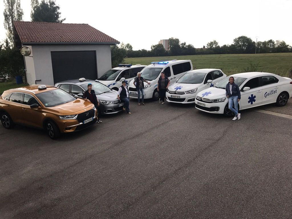 Cinq voitures garées devant un garage, avec des personnes debout à côté, sur un parking pavé.